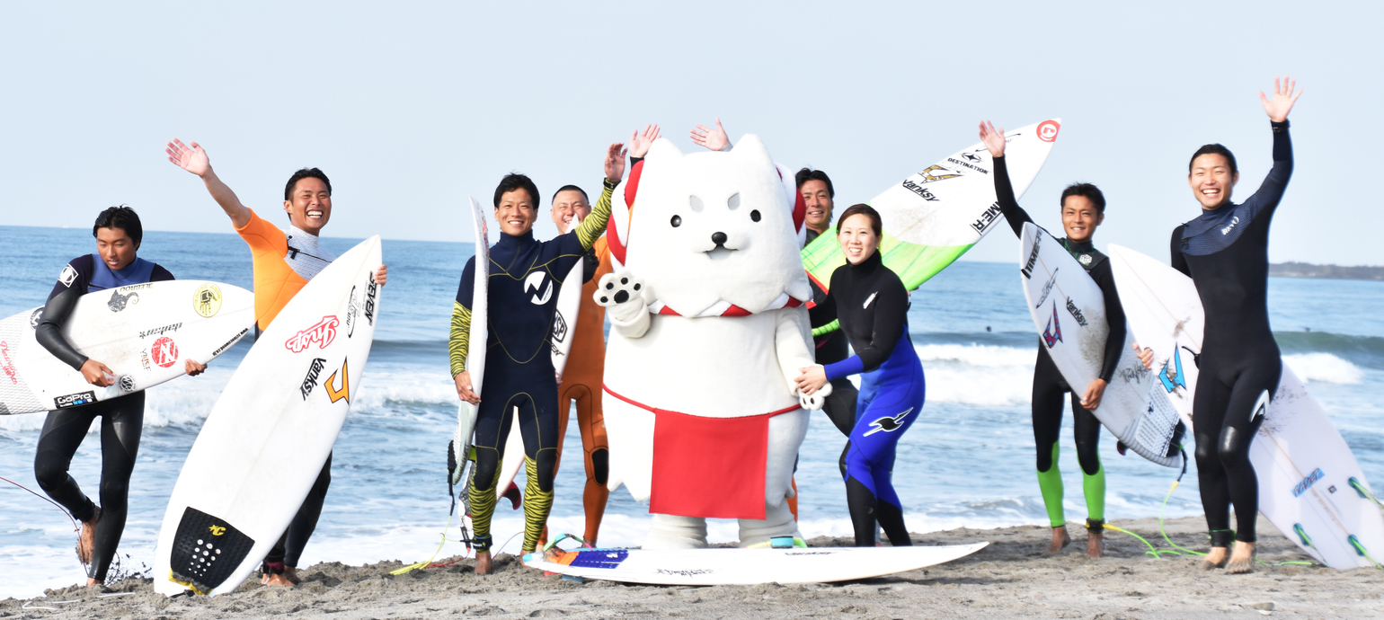 写真：海を背景にサーフィンボードを持ち笑顔の若者としっぺい